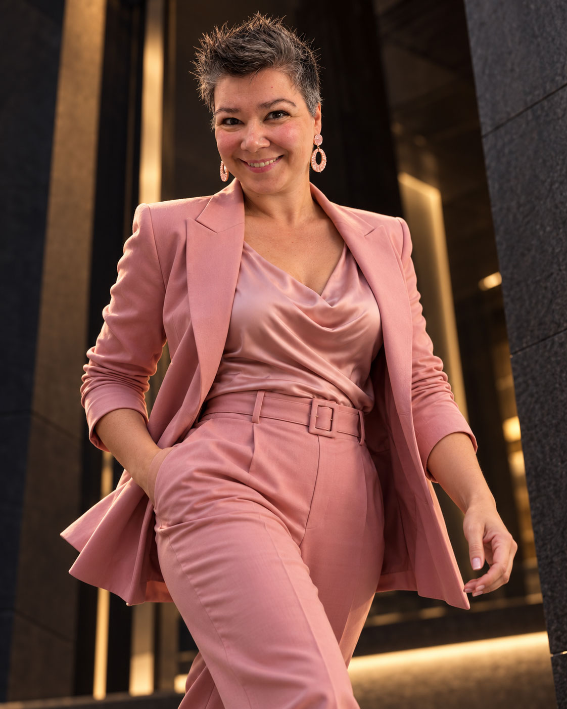 Marita im rosa Hosenanzug, in Bewegung vor dunkler Architektur